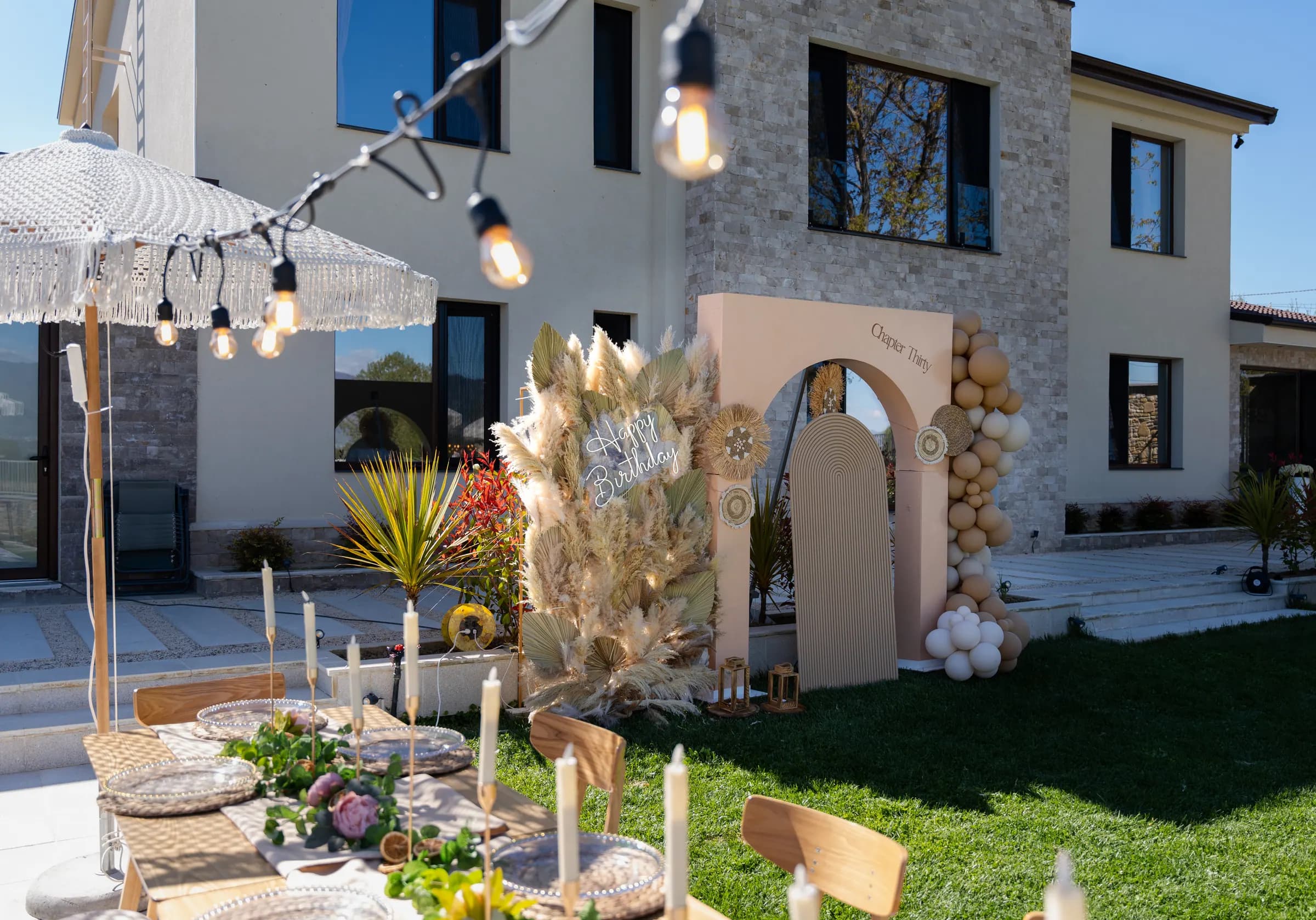 Tavolinë e gjatë festive në kopsht me pampas, qirinj dhe ombrella boho
